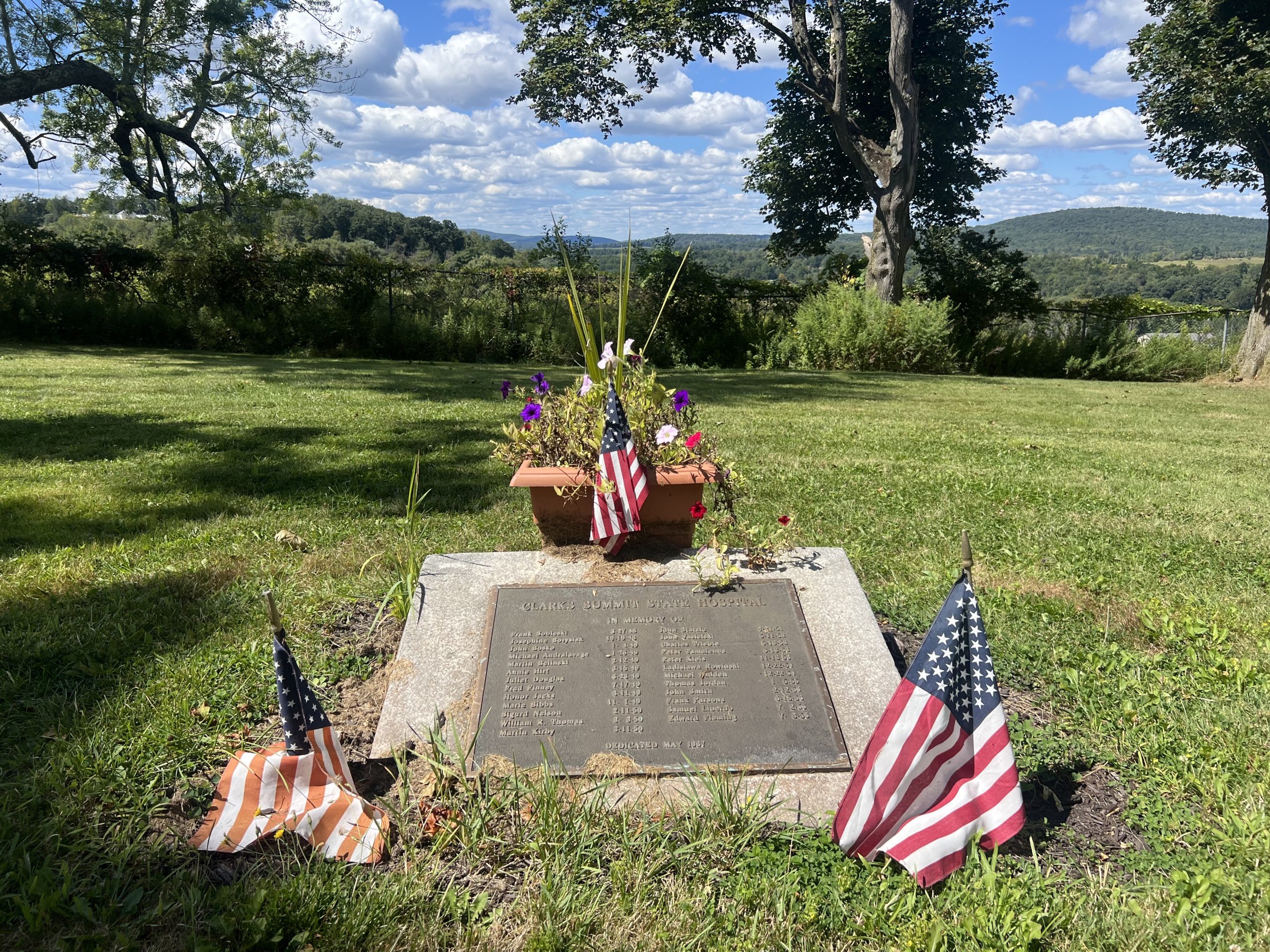 A memorial plaque on the ground, with American flags and flower on its edges. The plaque lists twenty-five names